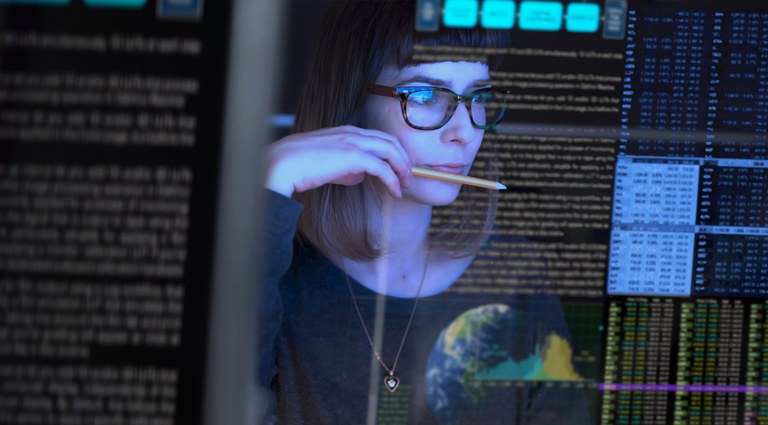 Foto com uma mulher olhando para várias telas de computador preenchidas com muitos dados