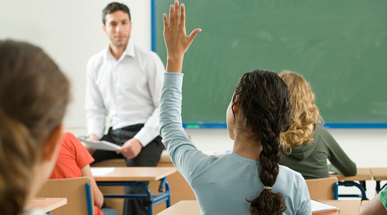 Foto com professor e alunos, em sala de aula. Uma aluna está com mão levantada, para fazer uma pergunta