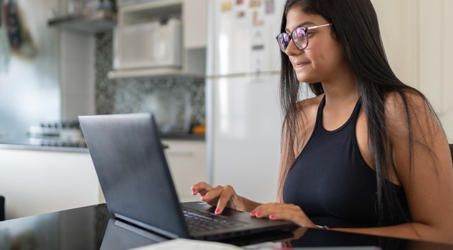 Moça jovem usando um notebook, o qual está em uma mesa na cozinha da casa