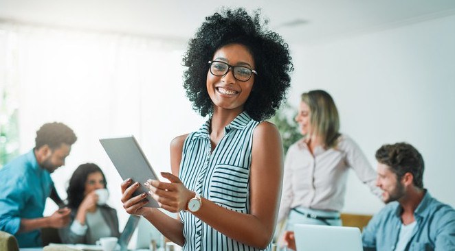 Imagem de uma mulher sorridente, em destaque, segurando um notebook, com pessoas ao fundo, como se estivessem em reunião, conversando