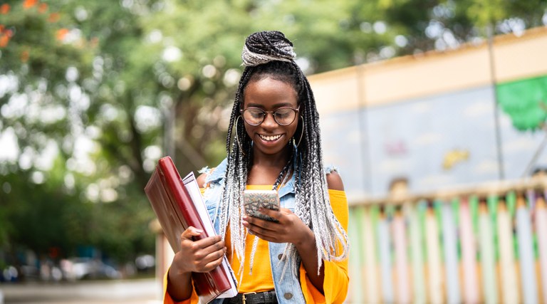 Imagem com jovem negra, sorridente, olhando para a tela do celular, o qual está em uma de suas mãos. Na outra mão, ela carrega papéis e uma pasta