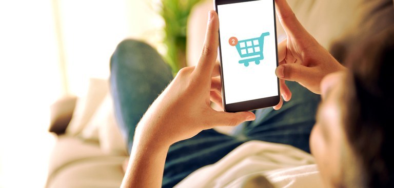 Uma pessoa segura um celular cuja tela exibe um ícone de um carrinho de compras online