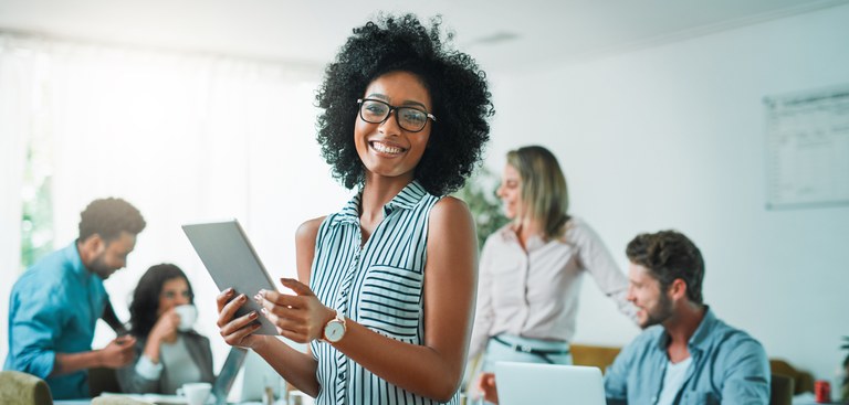 Mulher negra olha para frente e sorri enquanto setgura um tablet. Atrás dela, dois homens e duas mulheres conversam numa mesa com um notebook aberto