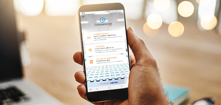 Imagem com uma mão segurando um celular. Na tela do celular, imagem do app Comprasnet Mobile e suas funcionalidades