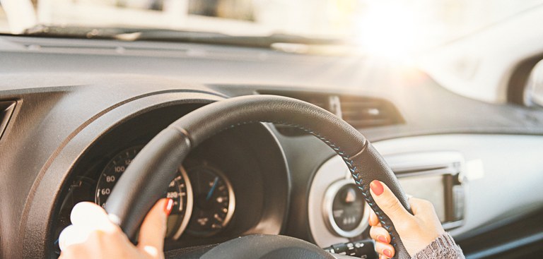 Carro visto por dentro. Mão feminina segura volante com brilho do sol ao lado.