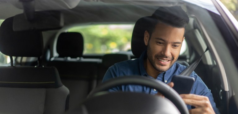 Foto com vista próxima do para-brisa de um carro. Há um homem no banco do motorista que sorri ao utilizar o celular.