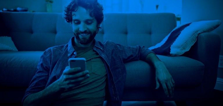 Um homem sorrindo, sentando no chão e encostado no sofá, na sala de casa, olhando para a tela do celular