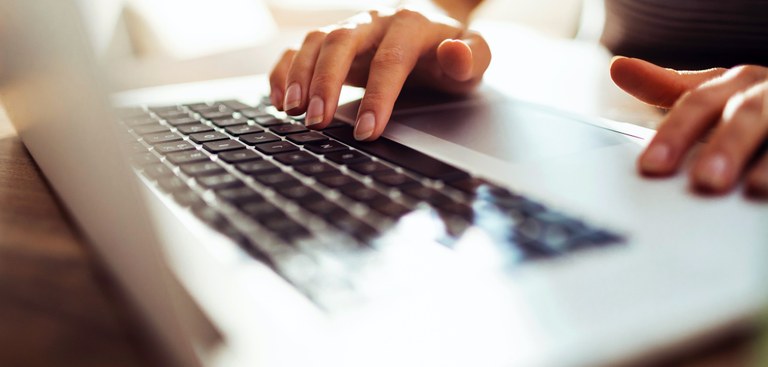 Foto em tons claros. Mãos femininas utilizam um notebook de cor prata, que é exibido lateralmente e de forma parcial, com a parte de trás do monitor desfocada e destaque para o teclado.