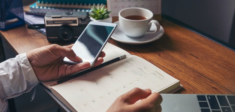 Foto de mesa de escritório com xícara de café, calendário, cadernos, notebook, livros e uma máquina fotográfica retrô. Mão feminina segura um celular apoiada sobre uma agenda, com um lápis apoiado.