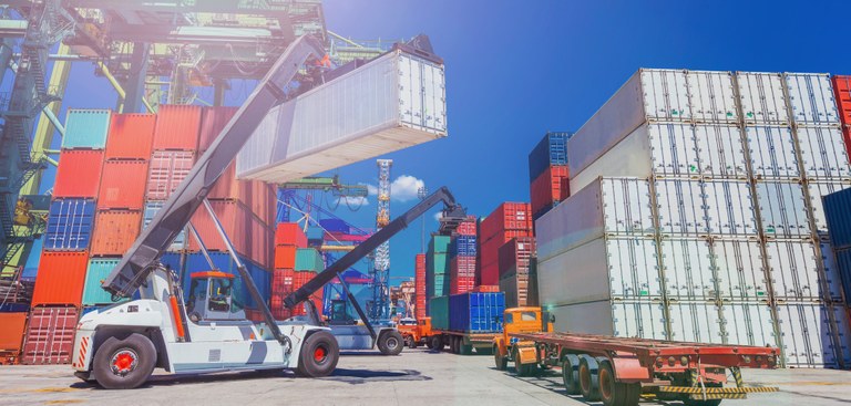 Foto de um pátio cheio de containers de variadas cores, um veículo com guindaste movimenta um container.