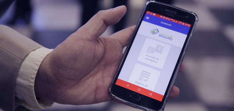 Fotografia de uma mão masculina exibindo um telefone celular cuja tela exibe o aplicativo do Embarque Mais Seguro