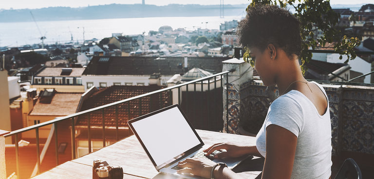 Mulher acessa um notebook em um terraço de onde se vê os telhados da cidade de Lisboa