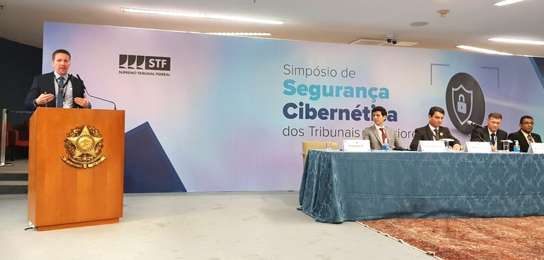 Tiago Iahn palestra no púlpito em Simpósio de Segurança Cibernética dos tribunais superiores, ao seu lado, quatro participantes assistem sentado à mesa.jpg