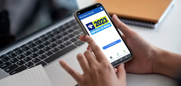 Mulher segura celular exibindo na tela o app Meu Imposto de Renda com imagem do IRPF 2023, ao fundo, um notebook e cadernos