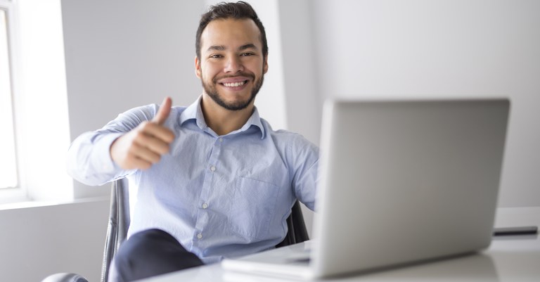 Homem pardo jovem sentado em frente a notebook fazendo gesto de legal com a mão direita.jpg