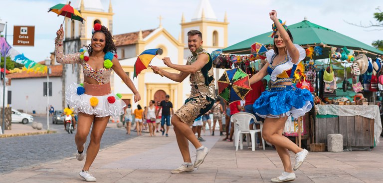 Foto de performance de dançarinos de frevo, duas mulheres e um homem numa praça, com feirinha e igreja ao fundo.jpg