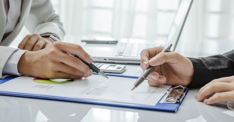 Mãos segurando canetas analisando um documento com gráficos, calculadora e notebook.jpg