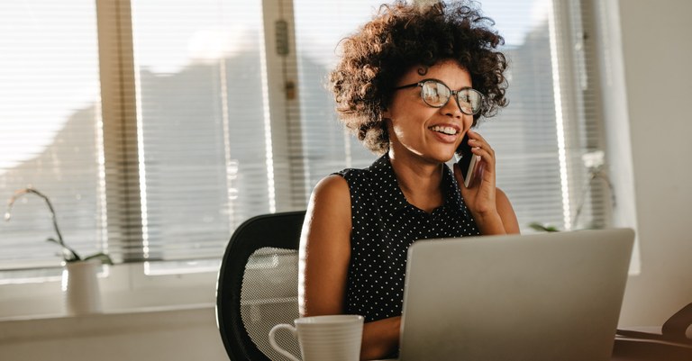 Mulher negra trabalhando em sua mesa com notebook e falando ao telefone.jpg