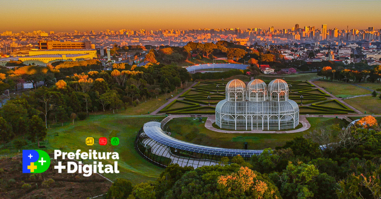 Vista área da cidade de Curitiba, com destaque para o Jardim Botânico Municipal. Sobre a imagem foi aplicada a marca do Prefeitura +Digital