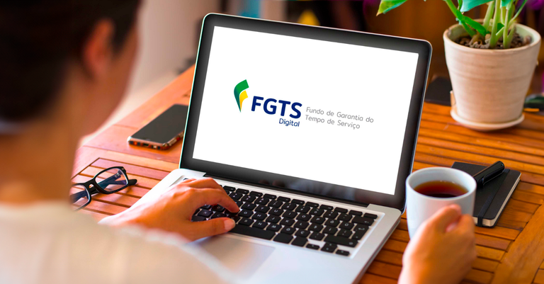 Foto de uma pessoa de costas, sentada numa mesa onde se vê um computador no qual aparece a marca do FGTS Digital na tela
