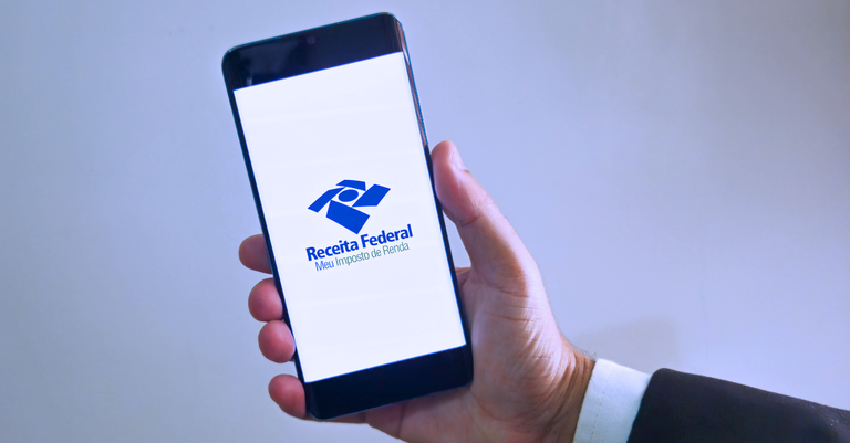 Fotografia de uma mão segurando um smartphone, cuja tela exibe a marca da Receita Federal e os dizeres 'Meu Imposto de Renda Fotografia de uma mão segurando um smartphone, cuja tela exibe a marca da Receita Federal e os dizeres 'Meu Imposto de Renda