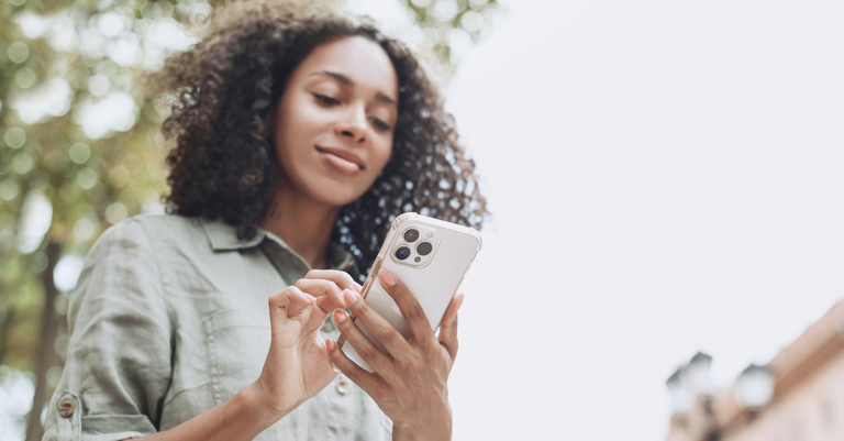 Mulher utilizando o celular. Ao fundo uma copa de árvore, indicando que ela está ao ar livre Mulher utilizando o celular. Ao fundo uma copa de árvore, indicando que ela está ao ar livre