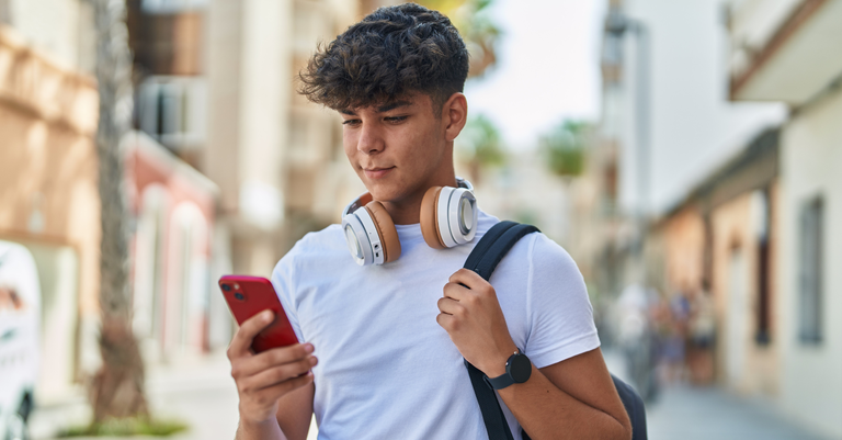 Fotografia de um estudante com mochila nas costas, fone de ouvido apoiado no pescoço e mexendo em um celular Fotografia de um estudante com mochila nas costas, fone de ouvido apoiado no pescoço e mexendo em um celular