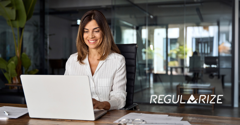 Mulher sorridente trabalha em um notebook em escritório. No lado direito da imagem está aplicada a marca do Portal Regularize